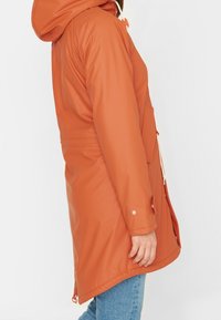 Veste orange imperméable avec capuche à cordon, poches latérales et coupe décontractée, au toucher lisse et avec un ourlet ajustable.