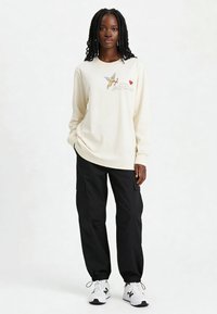 Chemise longue à manches longues de couleur crème avec un motif brodé représentant un chérubin et le texte  « je t'aime », associée à un pantalon cargo noir et des chaussures de sport blanches.