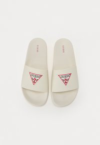 Sandale slide într-o nuanță crem, cu un logo Guess triunghiular roșu și alb. Material neted, cu o talpă plată și un breton lat. Design simplu.