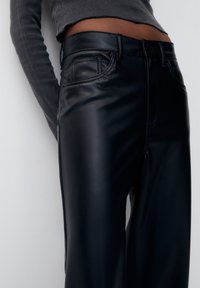 Pantalon en simili cuir noir avec une texture lisse, doté d'une taille mi-haute, de poches latérales et de détails de couture subtils.