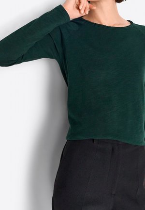 Osoba ubrana w ciemnozieloną bluzkę z długim rękawem z niestandardowym wykończeniem dekoltu i czarne spodnie, pozuje z uniesioną jedną ręką blisko twarzy.