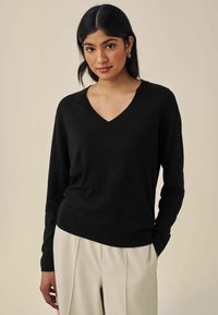 Pull noir à col en V avec des manches longues, fabriqué dans un tissu lisse. Porté sur un pantalon crème, présentant une coupe décontractée et des poignets côtelés.