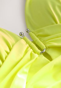 Neonově žlutý látkový top se sbíranou texturou a kovovými doplňky, vyrobený z hladkého, strečového materiálu a disponující unikátními designovými prvky.