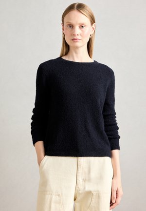 Woman with straight blonde hair wearing gold hoop earrings, black textured long-sleeve sweater, and cream high-waisted pants with hands in pockets.