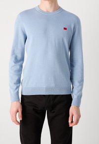Maglione lavorato a maglia di colore azzurro chiaro con maniche lunghe, polsini e orlo a coste, caratterizzato da un piccolo logo rosso sul petto. Texture liscia.