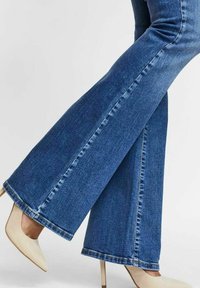 Jean évasé en denim bleu moyen avec une texture lisse, présentant une couture brute et des surpiqûres blanches, associé à des chaussures à talons beiges.
