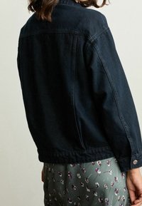 Chaqueta de mezclilla oscura con cuello tipo solapa, costuras visibles y puños abotonados, combinada con una falda de patrón floral en colores apagados.