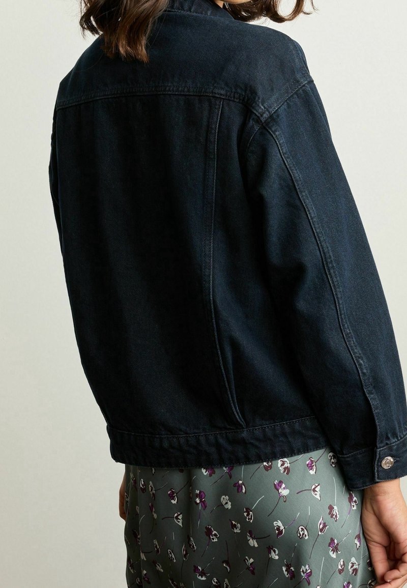 Chaqueta de mezclilla oscura con cuello tipo solapa, costuras visibles y puños abotonados, combinada con una falda de patrón floral en colores apagados.