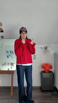 Red knitted sweater with ribbed hem, paired with dark blue flared jeans. Black beanie with white logo, holding a small object in one hand.