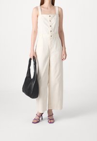 Ljusbeige denim overall med fyrkantig halsringning, framknappar och vida ben. Matchat med en svart axelremsväska och lila klackar med remmar.