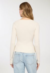 Top a maniche lunghe color crema con motivo a costine, silhouette aderente e vita leggermente a peplo, indossato con jeans denim azzurri.