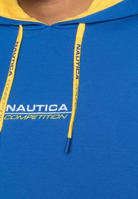 Blå huvtröja med gula accenter, med "Nautica Competition" broderat i vitt och gult. Inkluderar gula dragsnören med blå spetsar.