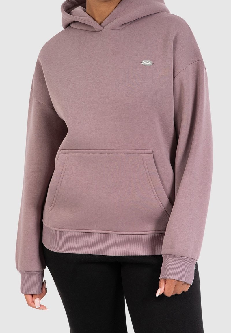 Femme portant un sweat à capuche mauve avec poche avant et petit logo, associé à un pantalon noir, visible des épaules jusqu’aux hanches.