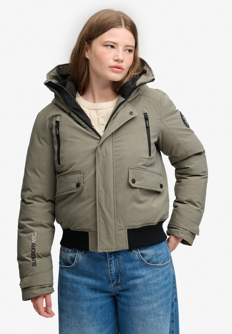 Giovane donna che indossa una giacca imbottita verde con zip e tasche e jeans blu, guarda leggermente di lato con le mani vicino ai fianchi.