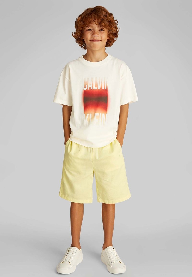 Lichtgekleurde katoenen T-shirt met rood-naar-zwart verloop "CALVIN KLEIN" tekst, gecombineerd met gele shorts en witte sneakers.