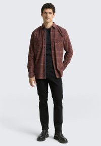 Chemise à carreaux bordeaux et marron foncé, associée à un t-shirt noir et un pantalon slim noir. Comprend deux poches à l'avant.