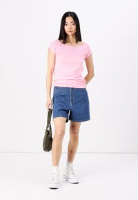 Top roz cu mânecă scurtă, pantaloni scurți din denim albastru, teniși înalți albi. Modelul ține o geantă mică verde, cu un fundal alb simplu.