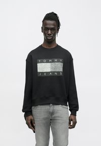 Hombre con rastas que lleva una sudadera negra Tommy Jeans y vaqueros gris claro, de pie contra un fondo blanco liso.