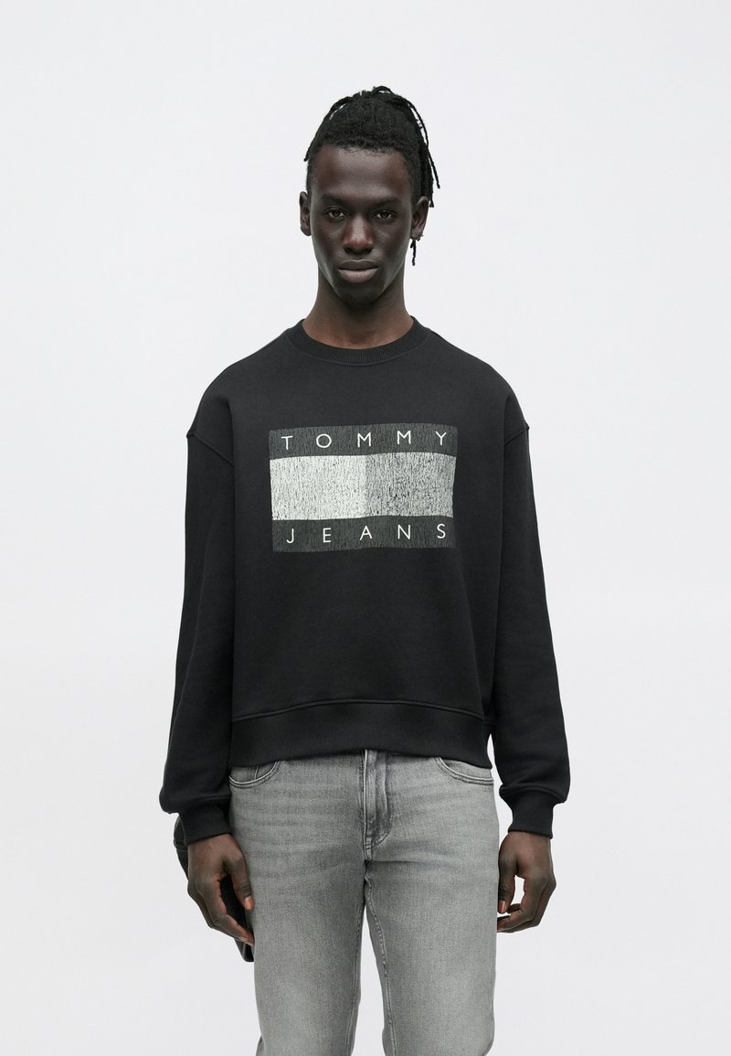 Hombre con rastas que lleva una sudadera negra Tommy Jeans y vaqueros gris claro, de pie contra un fondo blanco liso.