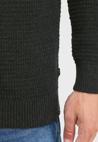 Svart strikket genser med teksturert mønster, ribbestrikkede mansjetter og nederkant. Synlig merketag på undersiden. Brukt sammen med blå denim jeans.
