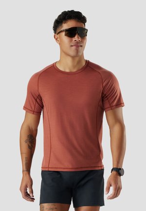 Mann trägt rostfarbenes Sport-T-Shirt, schwarze Shorts, schwarze Sonnenbrille und Smartwatch, steht mit neutralem Gesichtsausdruck vor einfachem Hintergrund.