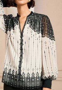 Svart och vit mönstrad blus med genomskinligt tyg, volangkrage, resårmanschetter och knapprad längs framsidan.