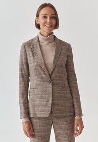Brūns šallīts žakete ar izceltām klajām, vienu pogu aizdari un divām priekšējām kabatām. Valkāta virs beiggai ribojuma turtleneck. Gluda tekstūra.