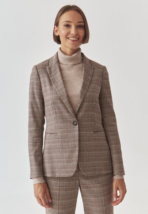 Hnědý kostkovaný blazer s špičatými klopami, jednořadovým zapínáním na knoflík a dvěma předními kapsami. Nosený přes béžový žebrovaný rolák. Hladká textura.