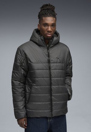 Giacca nera imbottita con cappuccio e chiusura zip, caratterizzata da un design a quilting orizzontale e tasche laterali, indossata sopra jeans blu.