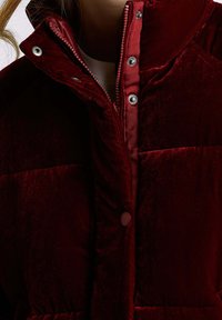 Bordeauxrode fluwelen pufferjas met een hoge kraag, drukknoopsluiting en gedetailleerde stiksels langs de rits en naden. Gestructureerde stof.