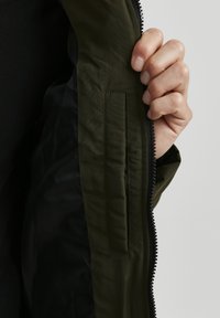 Olive green jacket with a smooth texture, featuring a zippered front and an interior pocket. The inside is black and subtly contrasting.