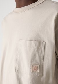 Ljusbeige långärmad skjorta i bomull med en framtill placerad ficka och en broderad logotapp. Rundad halsringning med subtila ribbad detaljer.