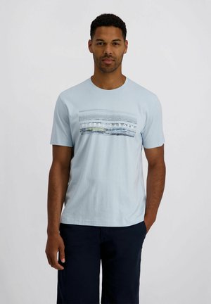 Mann trägt ein hellblaues T-Shirt mit abstrakten grauen Linien und Text, kombiniert mit dunkler Hose, steht vor einfarbigem Hintergrund.
