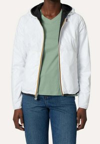 Vit zip-up jacka med huva, med svart foder och en gul accentdragkedja. Bärs över en grön V-ringad tröja och blå jeans.