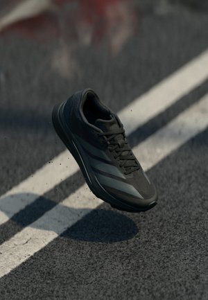 Czarne buty sportowe z teksturowaną siateczkową cholewką, z szarymi akcentami i amortyzowaną podeszwą, zawieszone w powietrzu nad chodnikiem.