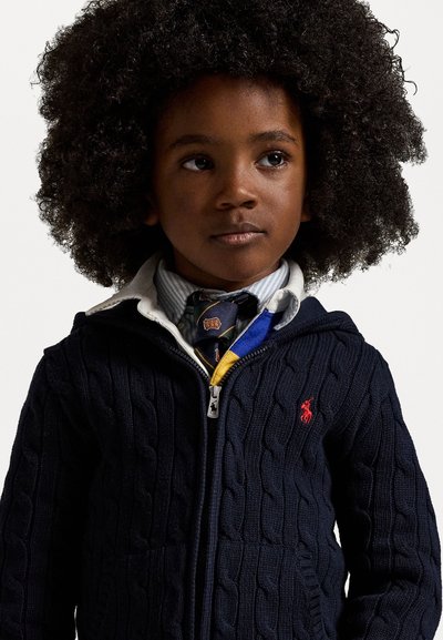 Jersey de punto en color navy con cremallera y logo rojo, superpuesto sobre una camisa de rayas azul y blanca y una corbata estampada. Textura suave, diseño casual.