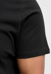 Juodas medvilninis marškinėliai trumpomis rankovėmis, lygia tekstūra ir matoma siuvimo linija aplink rankovių kraštą. Artimas vaizdas parodo audinio kritimą.