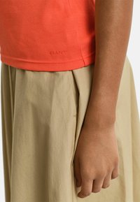 Orangenes Rippenshirt mit taillierter Passform, kombiniert mit einem beigen Faltenrock. Sichtbare Textur und Nähte verleihen dem Gesamtbild eine besondere Note.