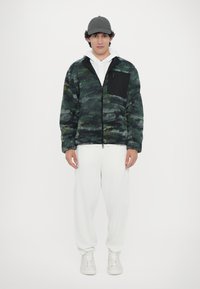 Camouflage-patterned fleece jacket in green and navy, with a black zippered chest pocket, paired with white pants and shoes.