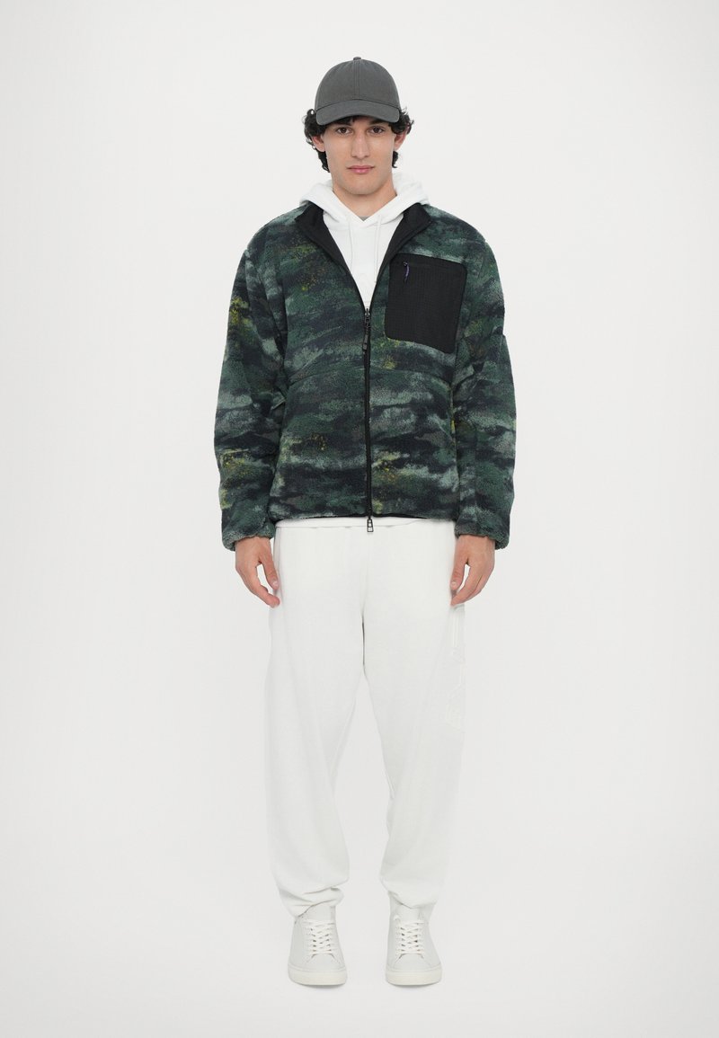 Camouflage-patterned fleece jacket in green and navy, with a black zippered chest pocket, paired with white pants and shoes.