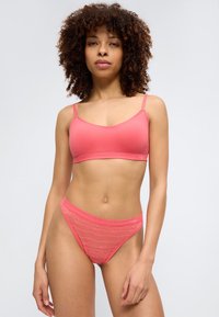 Femme aux cheveux bouclés portant un bralette corail rose et une culotte taille haute en dentelle assortie, debout devant un fond clair uni.