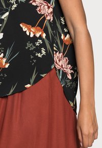 Blusa floreale nera con fiori arancioni e rosa, caratterizzata da un orlo curvo. Abbinata a una gonna di un solido colore ruggine. Tessuto dalla texture liscia.
