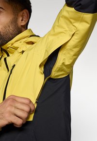 Vandeniui atsparus džemperis su geltona viršutine dalimi ir juoda apatine dalimi. Apima užtrauktuku uždaromas ventiliacijas ir kvėpuojančią pamušalą patogumui.