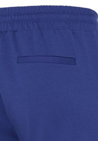 Gros plan d’un pantalon de survêtement à taille élastique bleue avec une seule poche passepoilée arrière du côté droit.