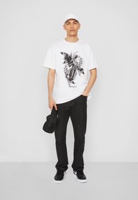 Hvid bomuld t-shirt med sort kunstværk og tekst i midten, parret med sorte denim-bukser og sorte sneakers, mens han/hun holder en sort kasket.