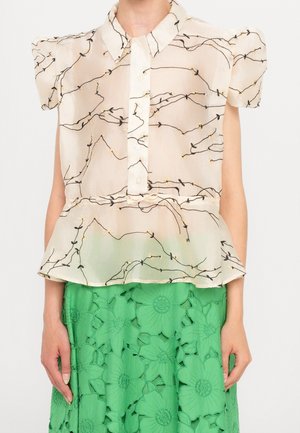 Blouse beige transparente avec imprimé vigne noire, manches courtes bouffantes, boutons sur le devant, associée à une jupe verte fleurie avec découpes.