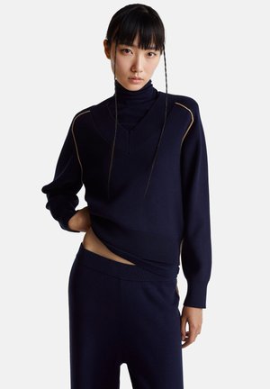 Námořnický pletený svetr s vysokým límcem a širokým výstřihem do tvaru V, s žebrovanými manžetami a lemem. Ozdobený tenkým béžovým lemem.