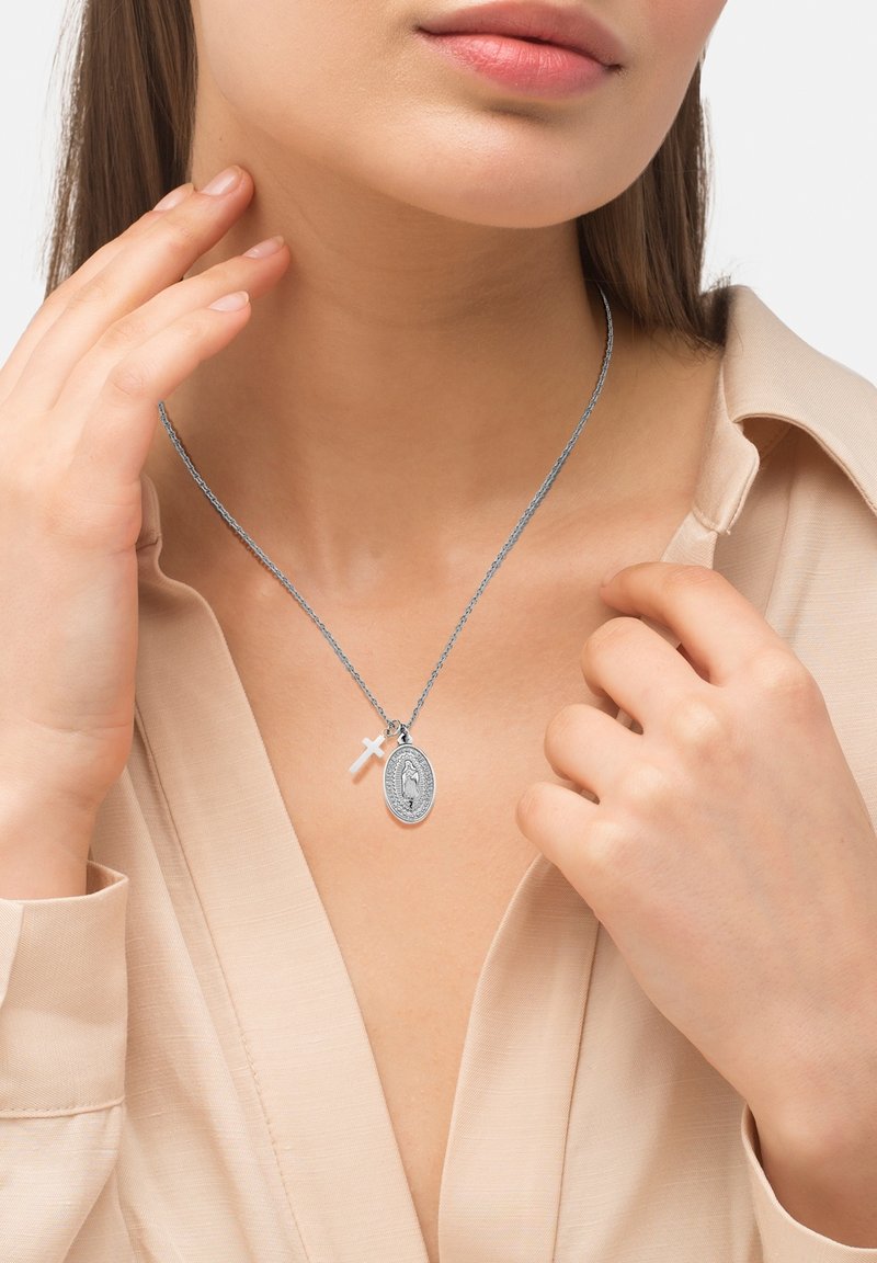 Femme portant une chemise beige touchant un collier en argent avec une croix et un pendentif religieux ovale autour du cou sur fond blanc.