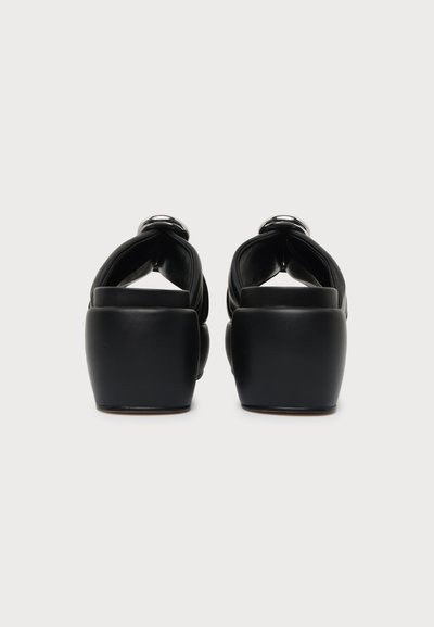 Paire de sandales noires à talons blocs vues de dos, avec de larges brides et des boucles argentées, sur fond blanc uni.