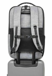 Sac à dos gris avec bretelles rembourrées et boucle de poitrine attachée à la poignée rétractable d'une valise rigide argentée sur fond blanc.